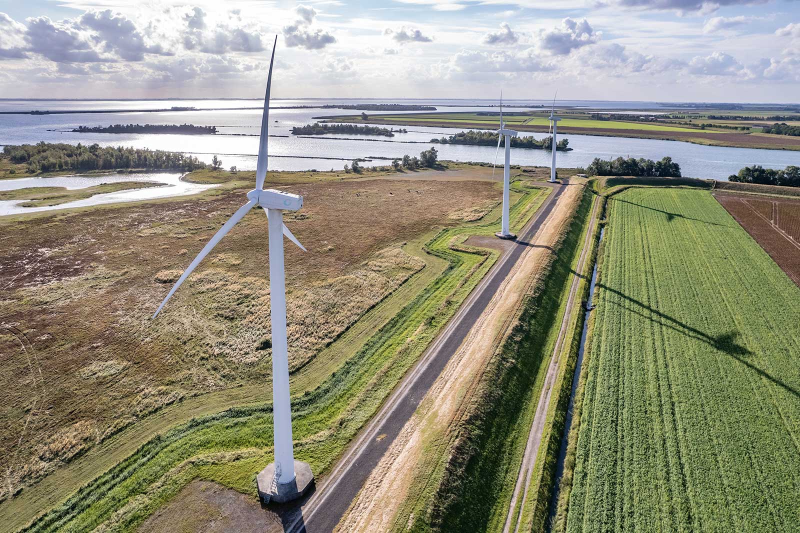 Three large wind turbines stand beside a winding road, with lush green fields and a shimmering waterway in the background.
