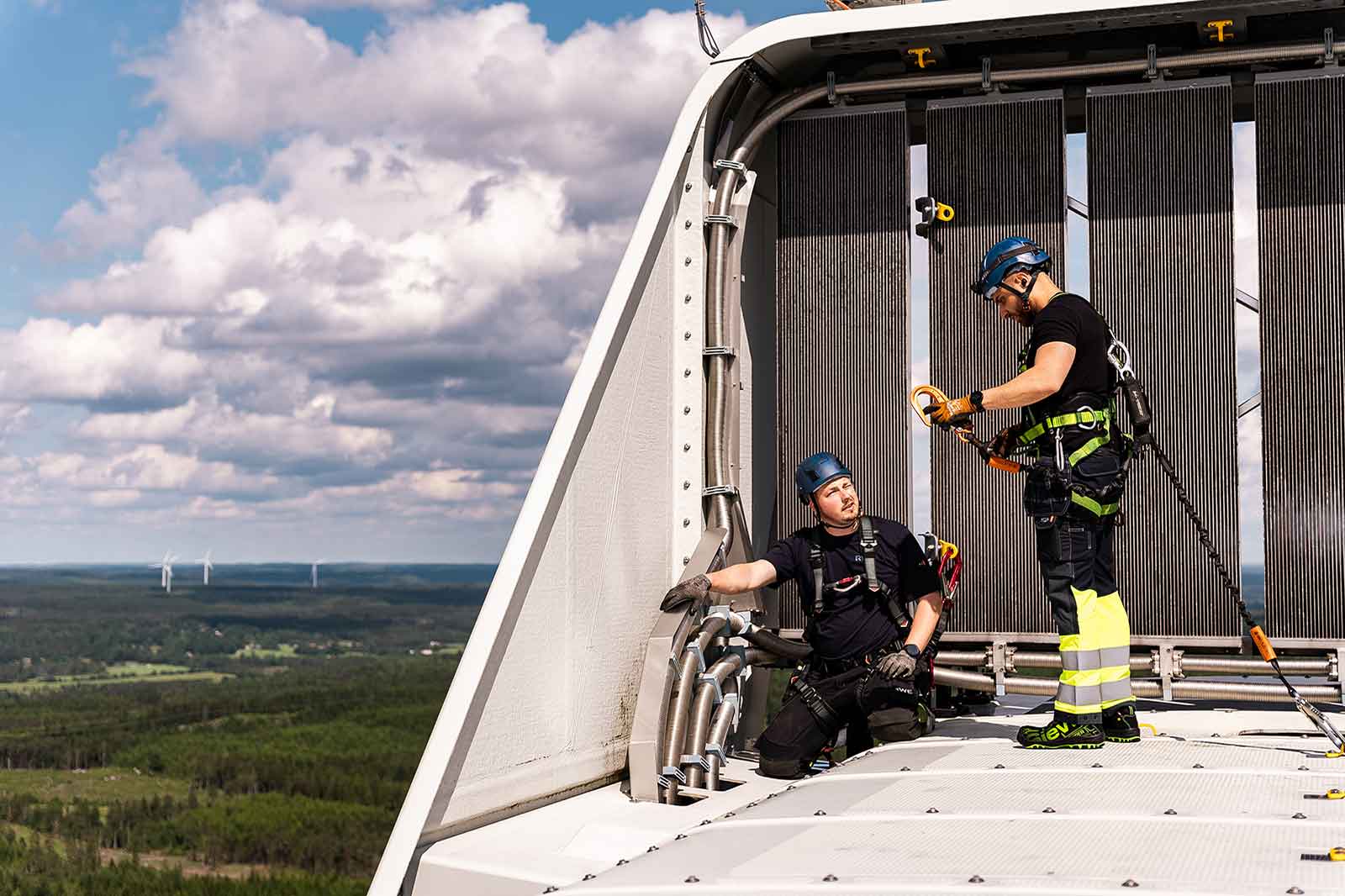 Two technicians work on top of a wind turbine, equipped with safety gear and harnesses against a backdrop of green fields and clouds.